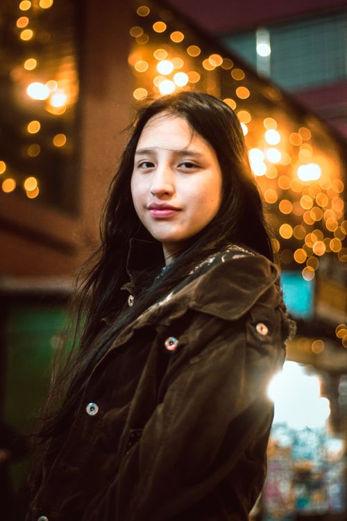 Free Young woman in a brown jacket standing against a cityscape with bokeh lights in the background. Stock Photo