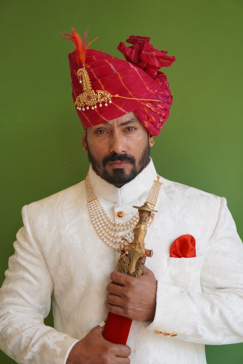 Free A man in a white suit and red turban posing with a sword against a green background. Stock Photo