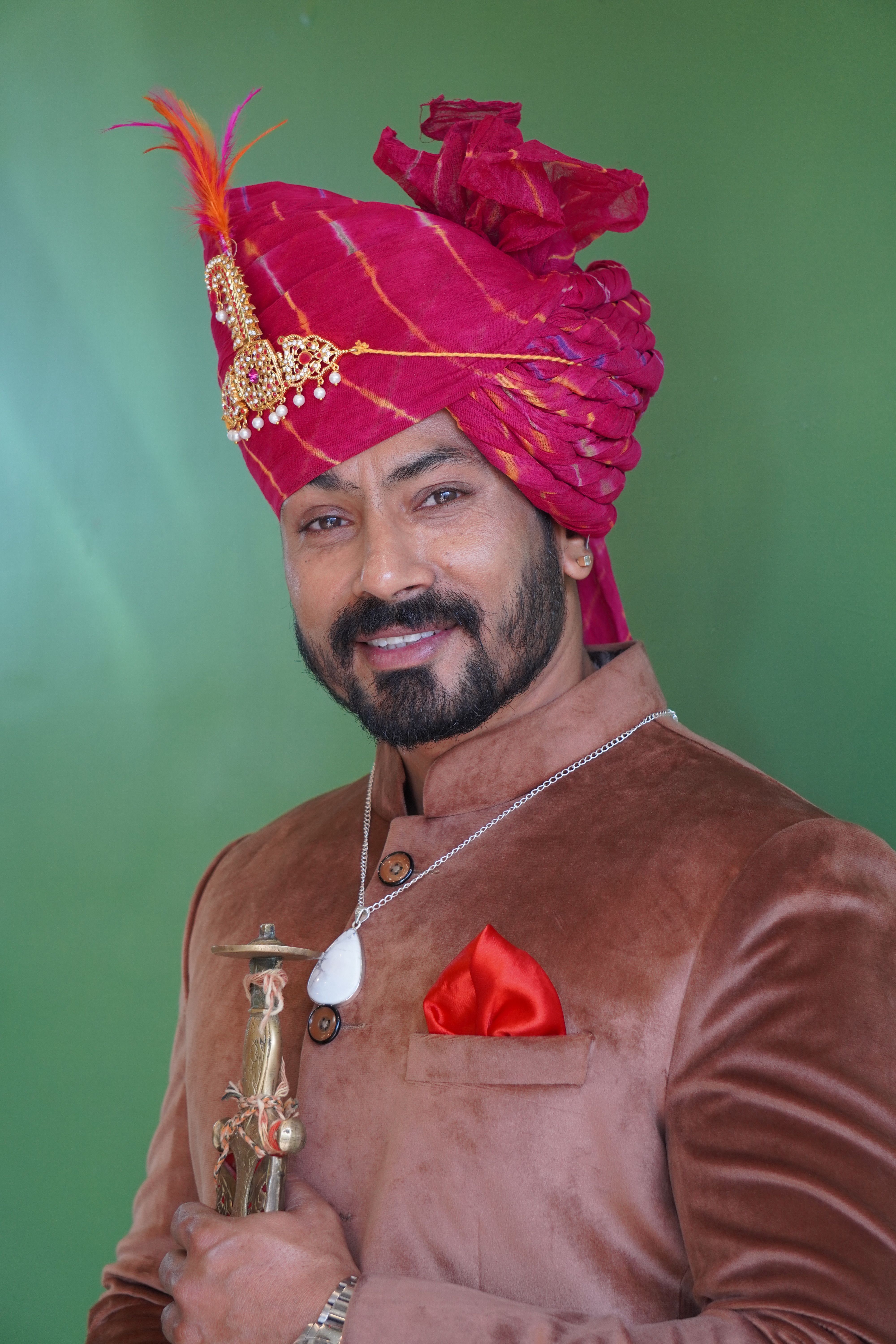 Portrait of an elegant man in traditional clothing and pink turban holding a sword on a green background.