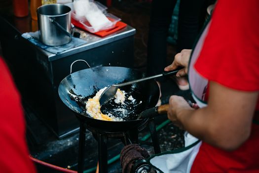 Close-up of a person cooking fried eggs in a wok at a lively Bangkok street market.