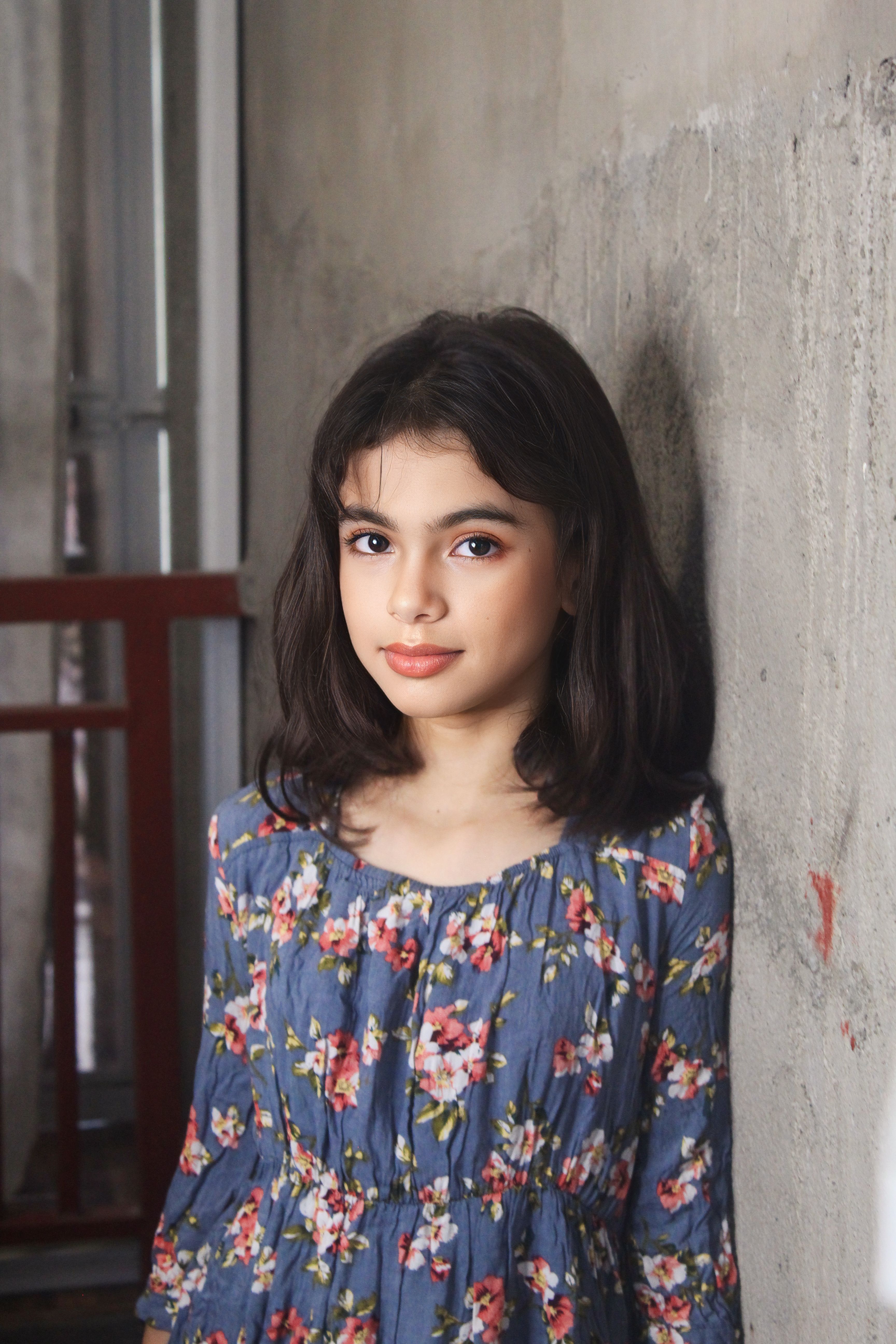 Portrait of a young girl with black hair in a floral dress leaning against an indoor wall.