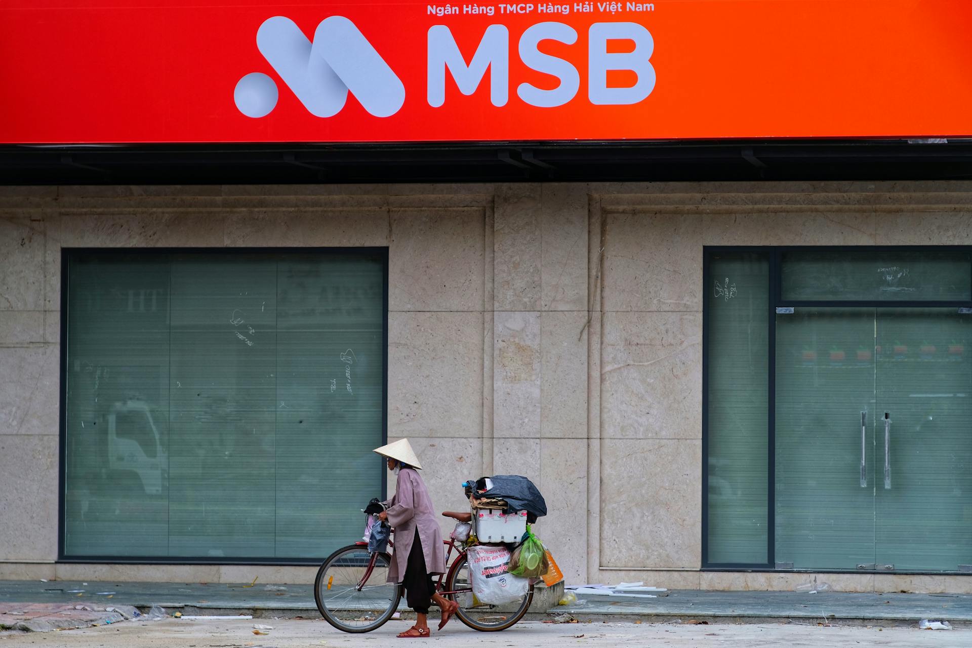 A traditional Vietnamese street vendor passes by a modern bank facade in a vibrant urban setting.