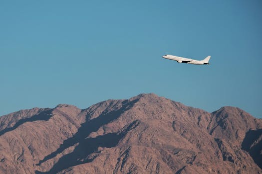 A white passenger airplane flies above rugged mountains under a clear blue sky, capturing the essence of travel.