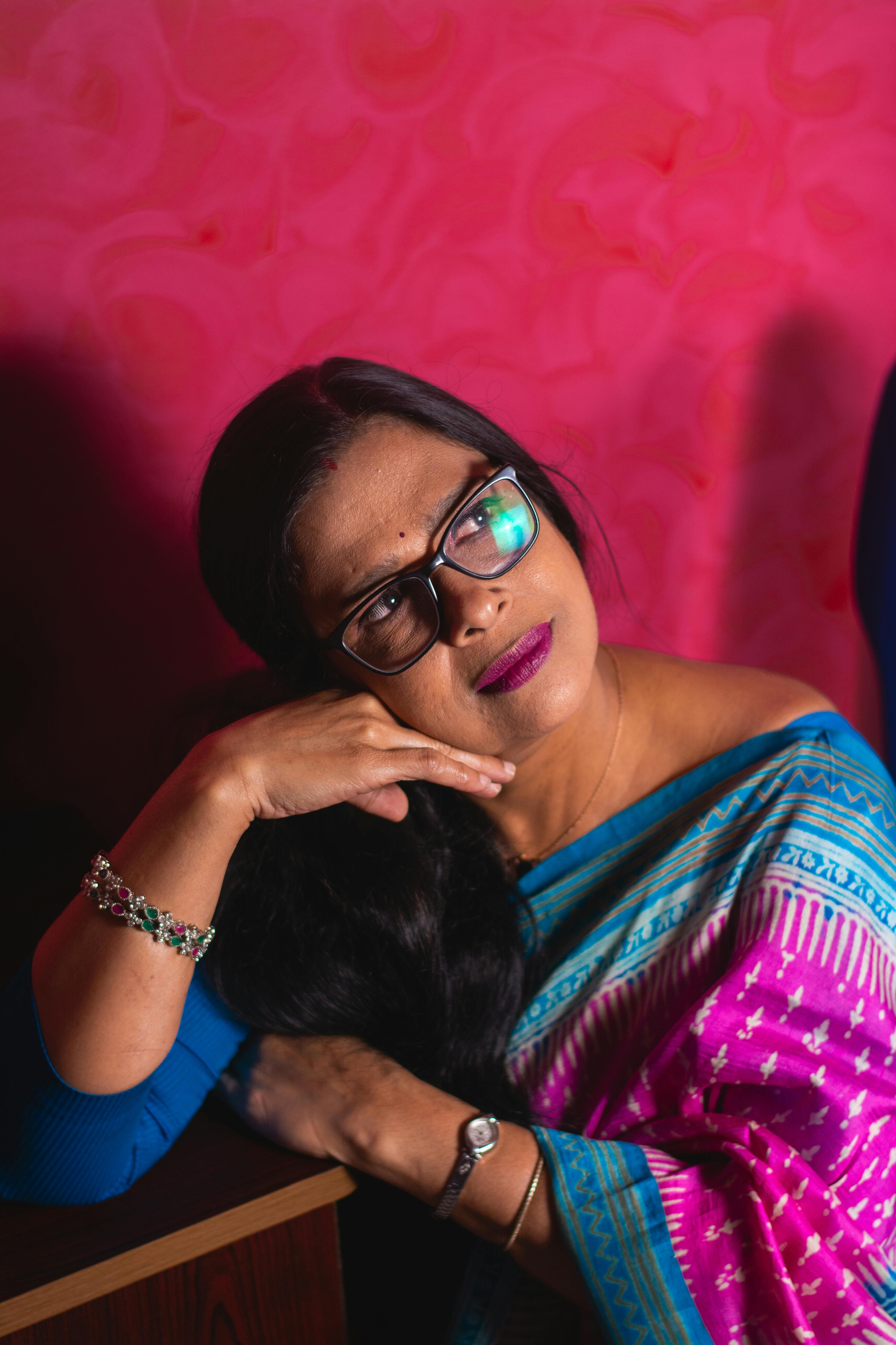 Contemplative Indian woman with eyeglasses in traditional sari. Vibrant pink background.
