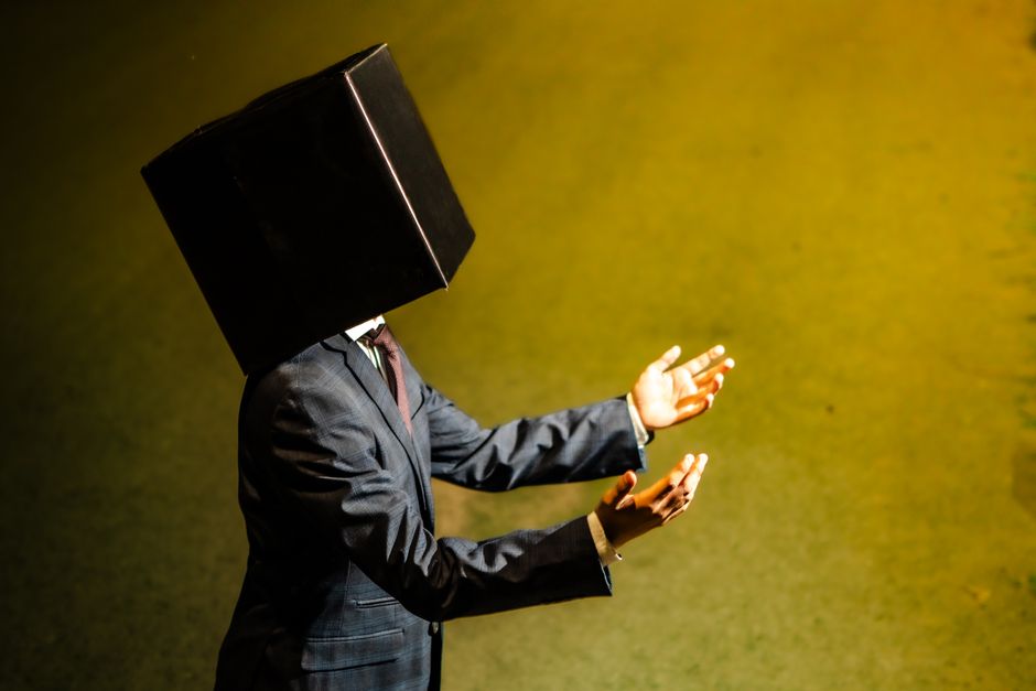 Person in suit with a black box on their head, arms raised, on a yellow-lit background.