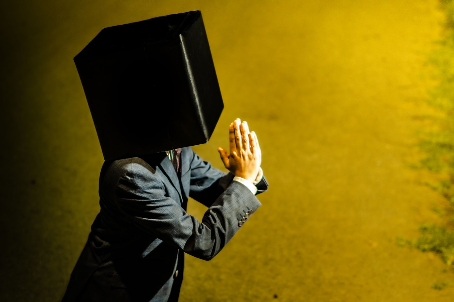 Conceptual image of a man in a suit with a black cube on his head praying on a yellow lit background.