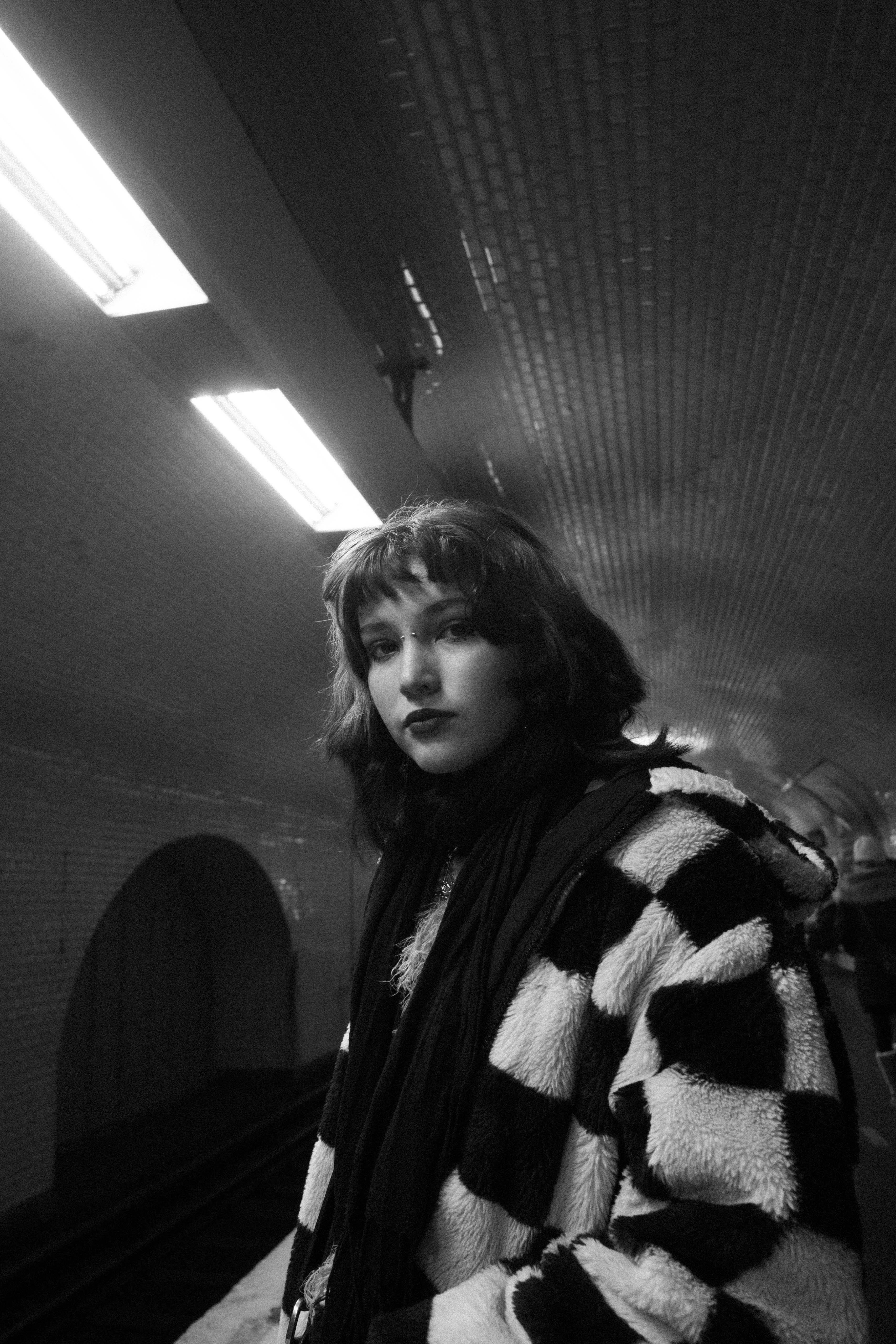 Fashionable woman in checkered coat, standing in a subway tunnel, captured in black and white.