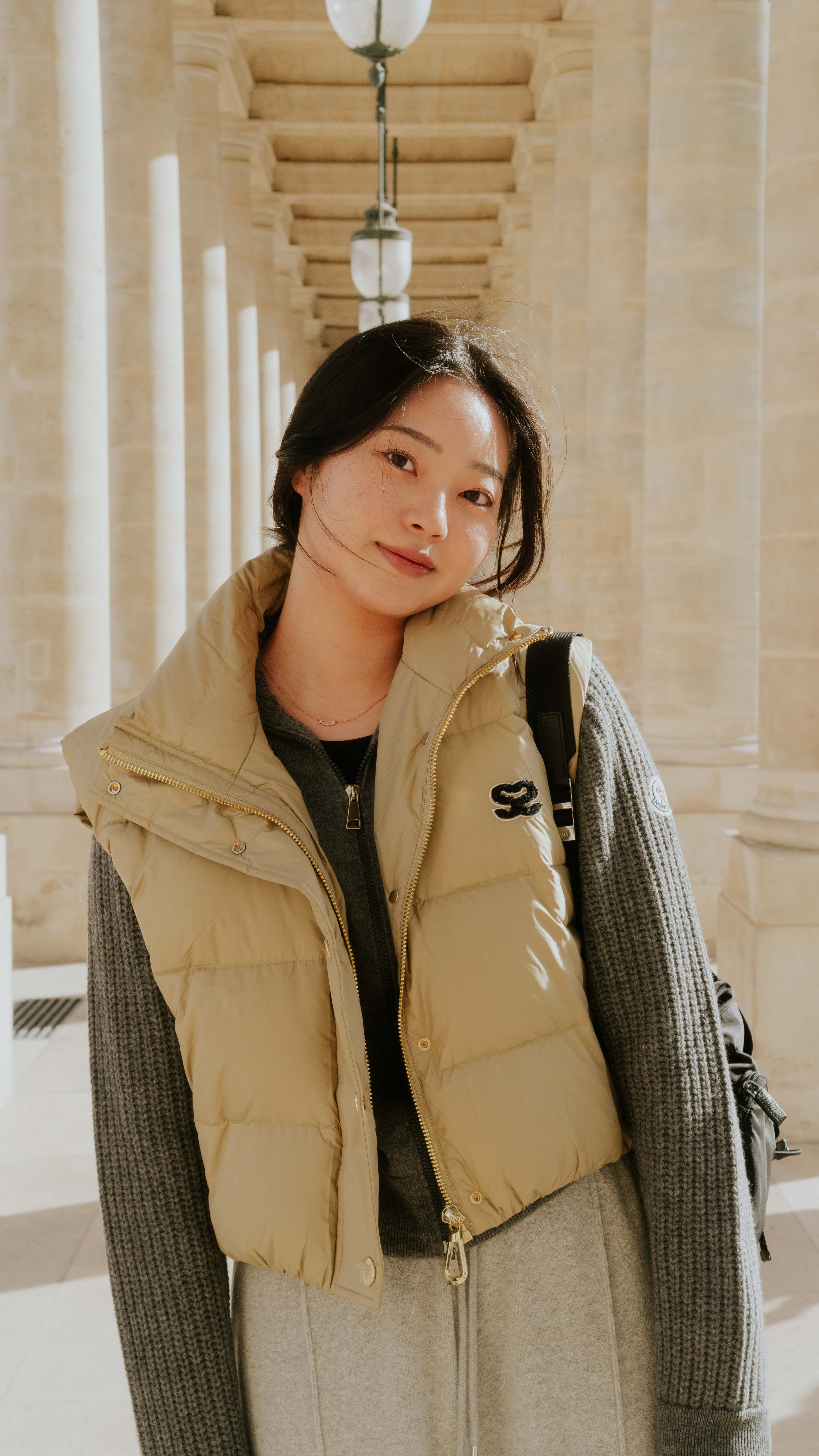A fashionable young woman posing in a quilted vest under architectural arches outdoors.