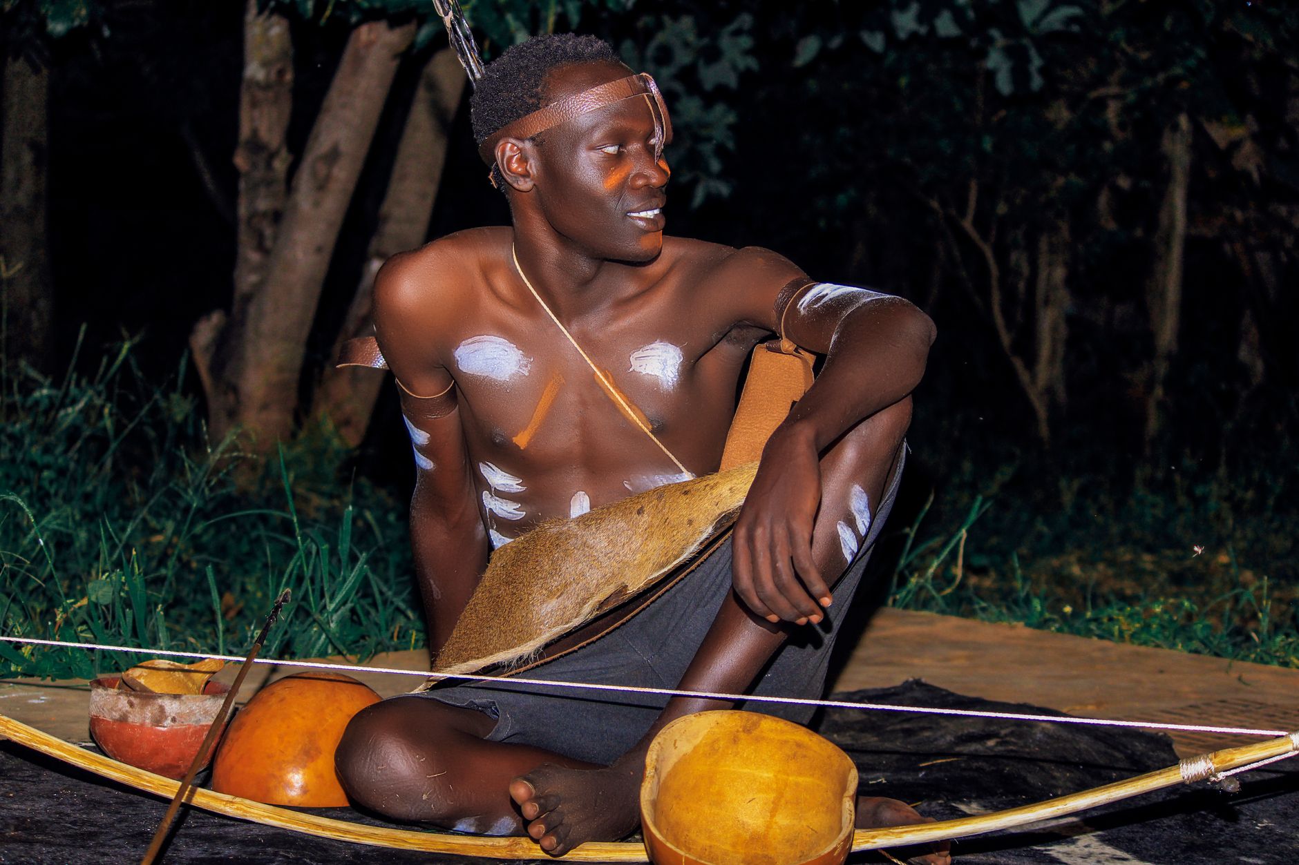 A young African man sitting outdoors with traditional tribal body paint and accessories at night in Uganda.