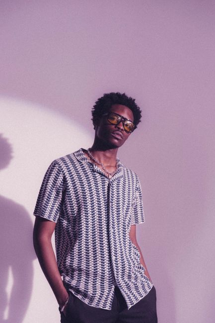 African man with curly hair and glasses, stylishly posing in a patterned shirt.