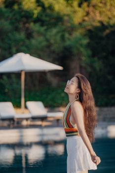 Smiling woman in summer attire enjoying the sun by a poolside with sunbeds in the background.