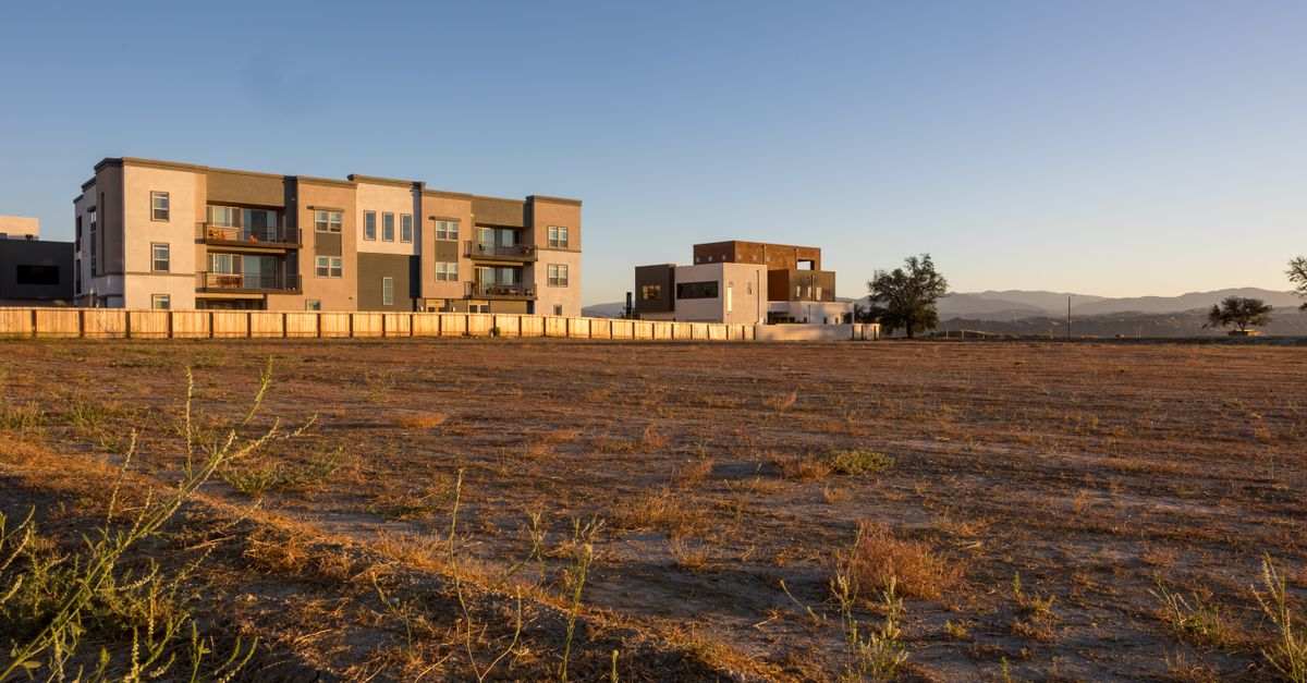Contemporary apartment blocks with ample open field space, framed by distant mountains under a clear blue sky.