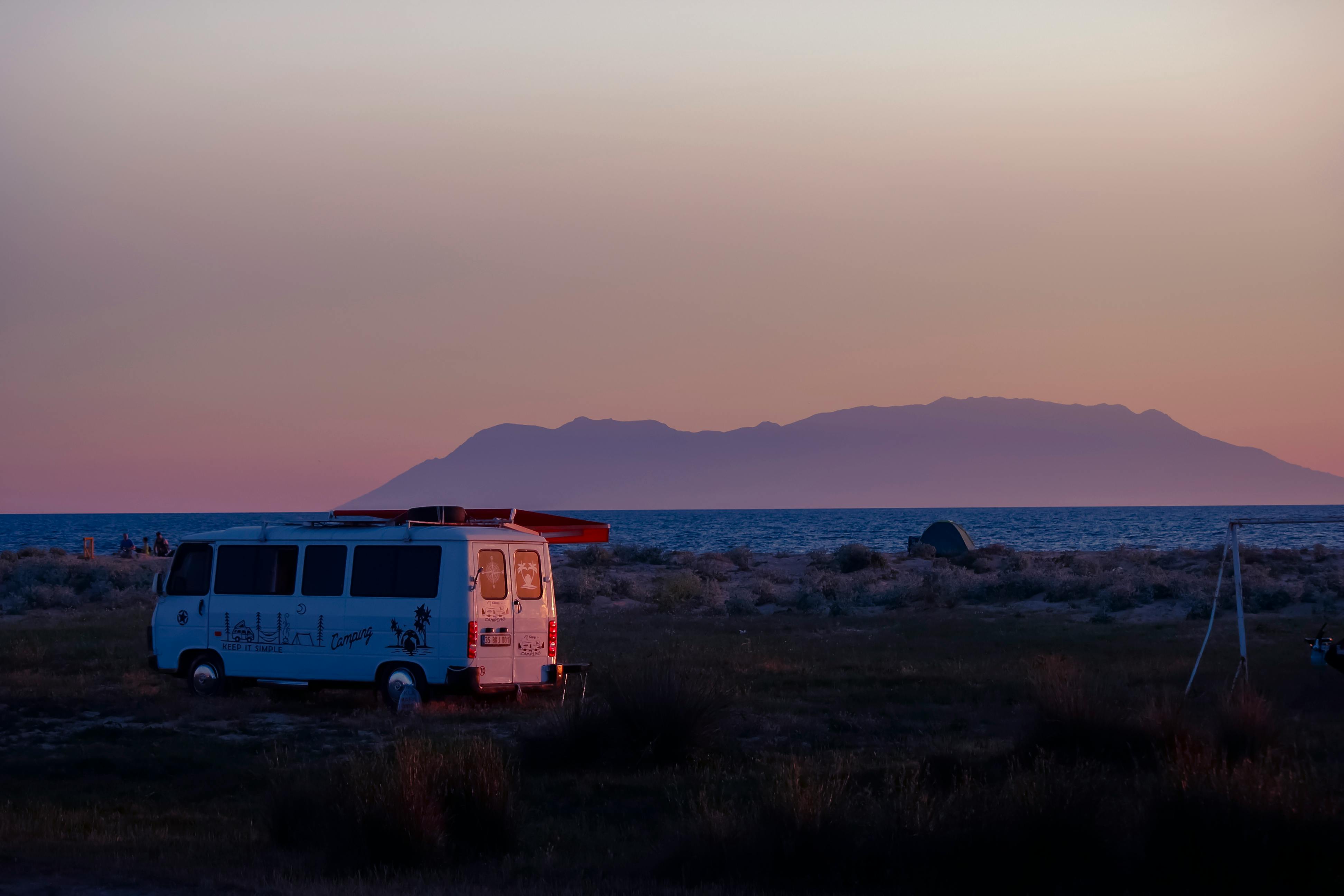 A tranquil sunset view featuring a van parked by the sea with mountains in the background.
