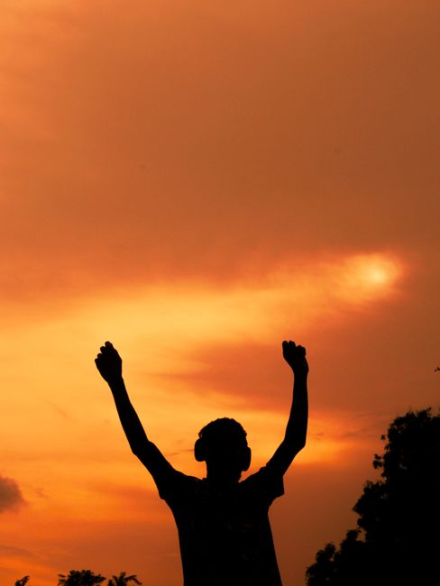 A silhouette of a man with headphones raised arms against a vibrant sunset sky.