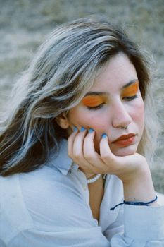 Close-up portrait of a woman with orange eye makeup, blue nails, and dyed hair, resting her head on her hand.