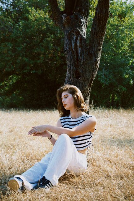 Woman with brown hair sitting under a tree in a sunny meadow, wearing striped top and white pants.
