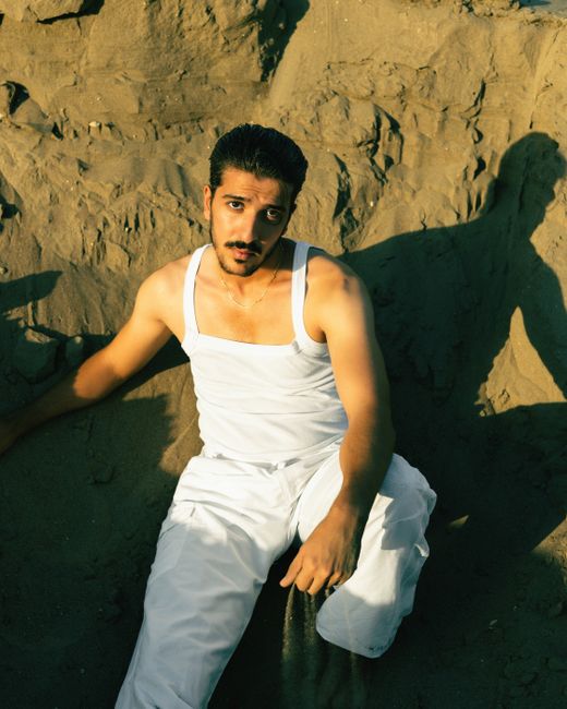 A man in a white tank top sits on a sandy beach, embracing the sun's warmth in Iraq.