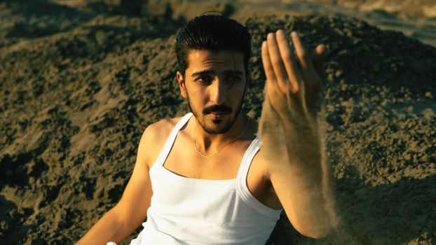 A man in a white tank top sitting on a sunny sandy beach, enjoying the warm weather.