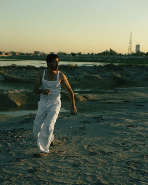 Free A man in white clothes runs on a sandy beach during sunset, with a city skyline in the background. Stock Photo