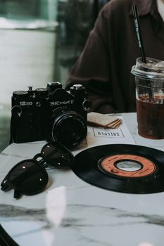 A vintage camera, vinyl record, and iced drink on a marble table create a nostalgic vibe.