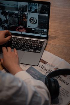 A person working on a laptop placed on a wooden desk with a newspaper and headphones nearby.