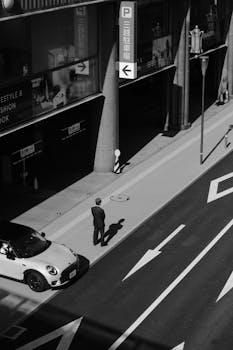 A man walks on a modern city street alongside a parked car in black and white.
