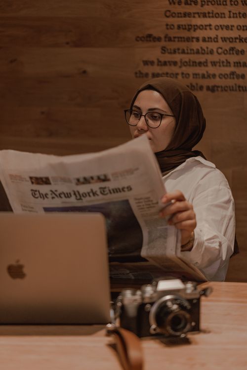 Free Muslim woman in hijab reading a newspaper at a café, laptop and camera on table. Stock Photo