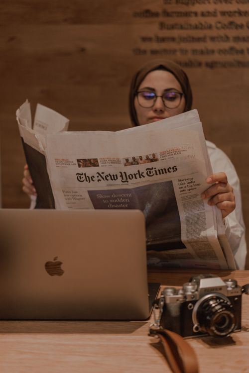 Free A Muslim woman in hijab reads a newspaper at a coffee shop, alongside a laptop and camera. Stock Photo