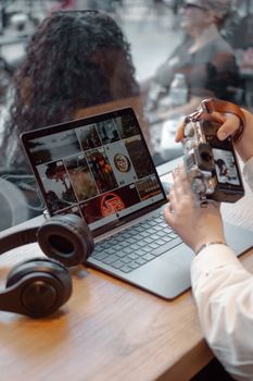 A woman using a laptop and vintage camera in a cozy café with headphones nearby.