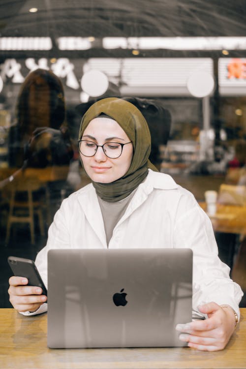 Free Middle Eastern woman multitasking with a laptop and smartphone at a cafe. Remote work concept. Stock Photo
