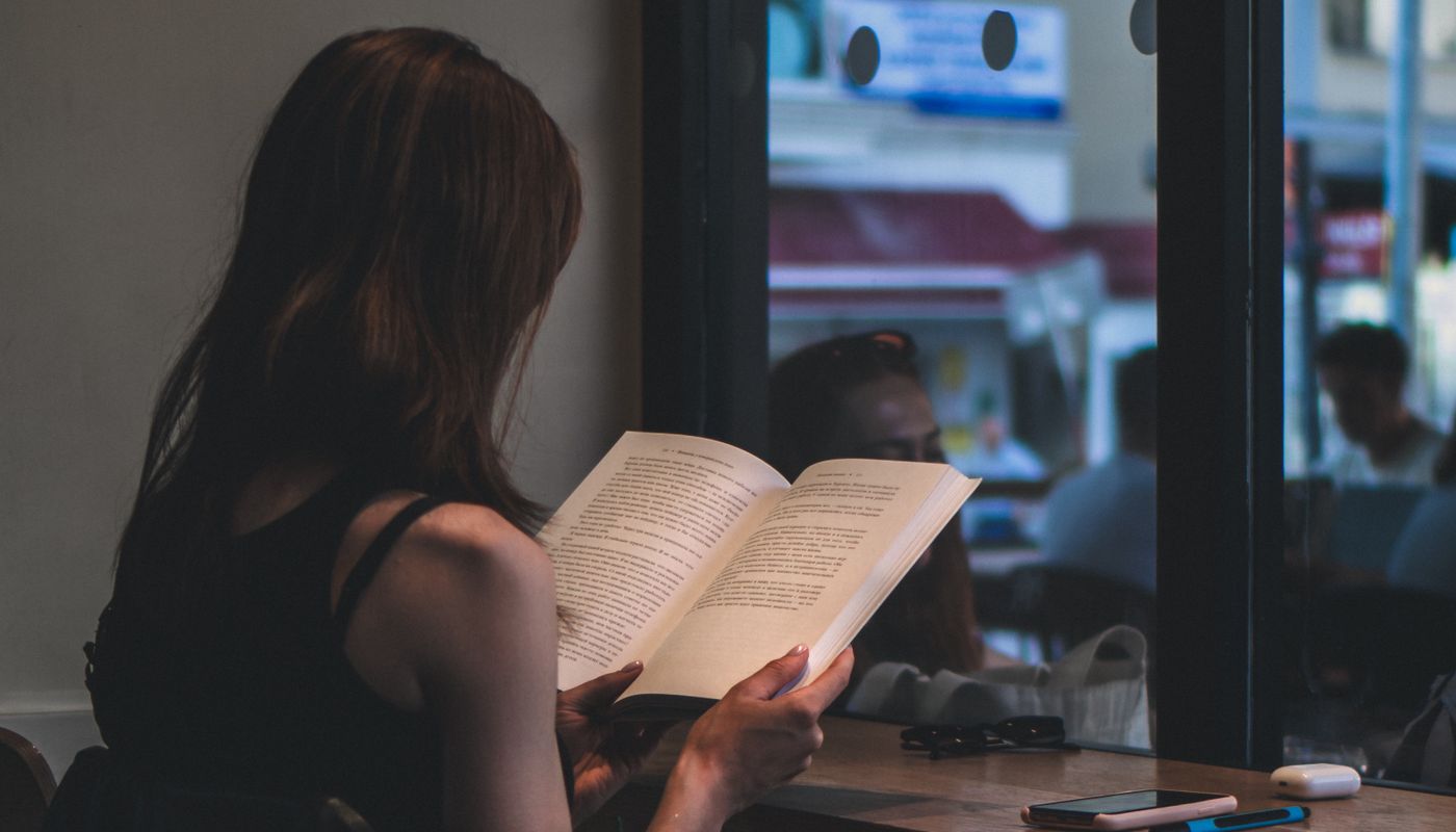 A person absorbed in a book beside a cafe window, a phone lying face-down on the table