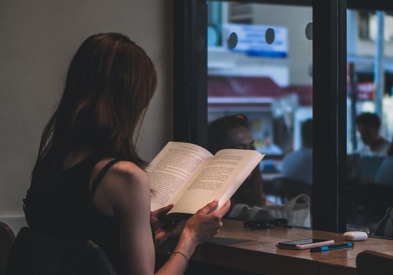 A person absorbed in a book beside a cafe window, a phone lying face-down on the table