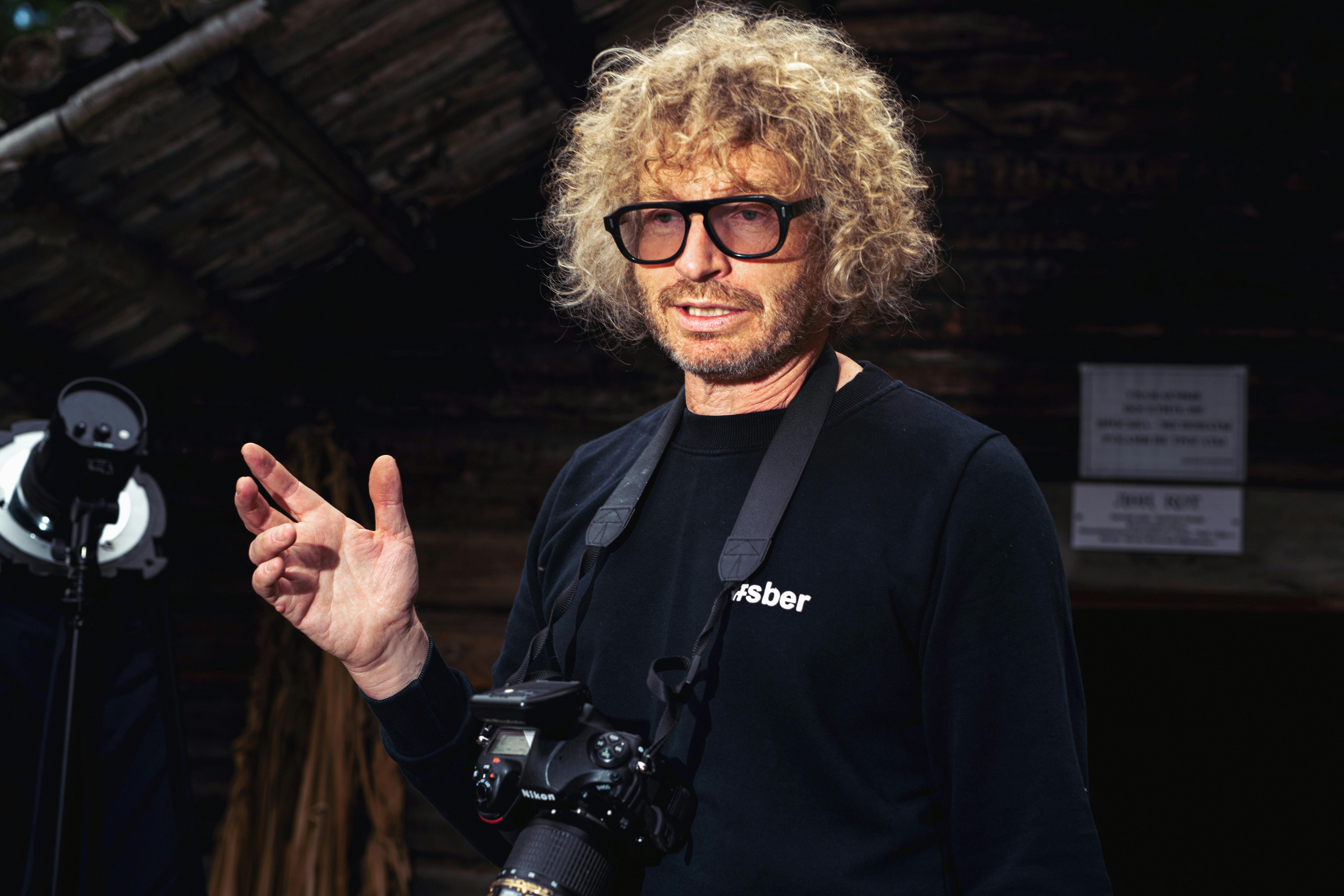 A photographer with curly blonde hair and eyeglasses holding a camera, standing outdoors.