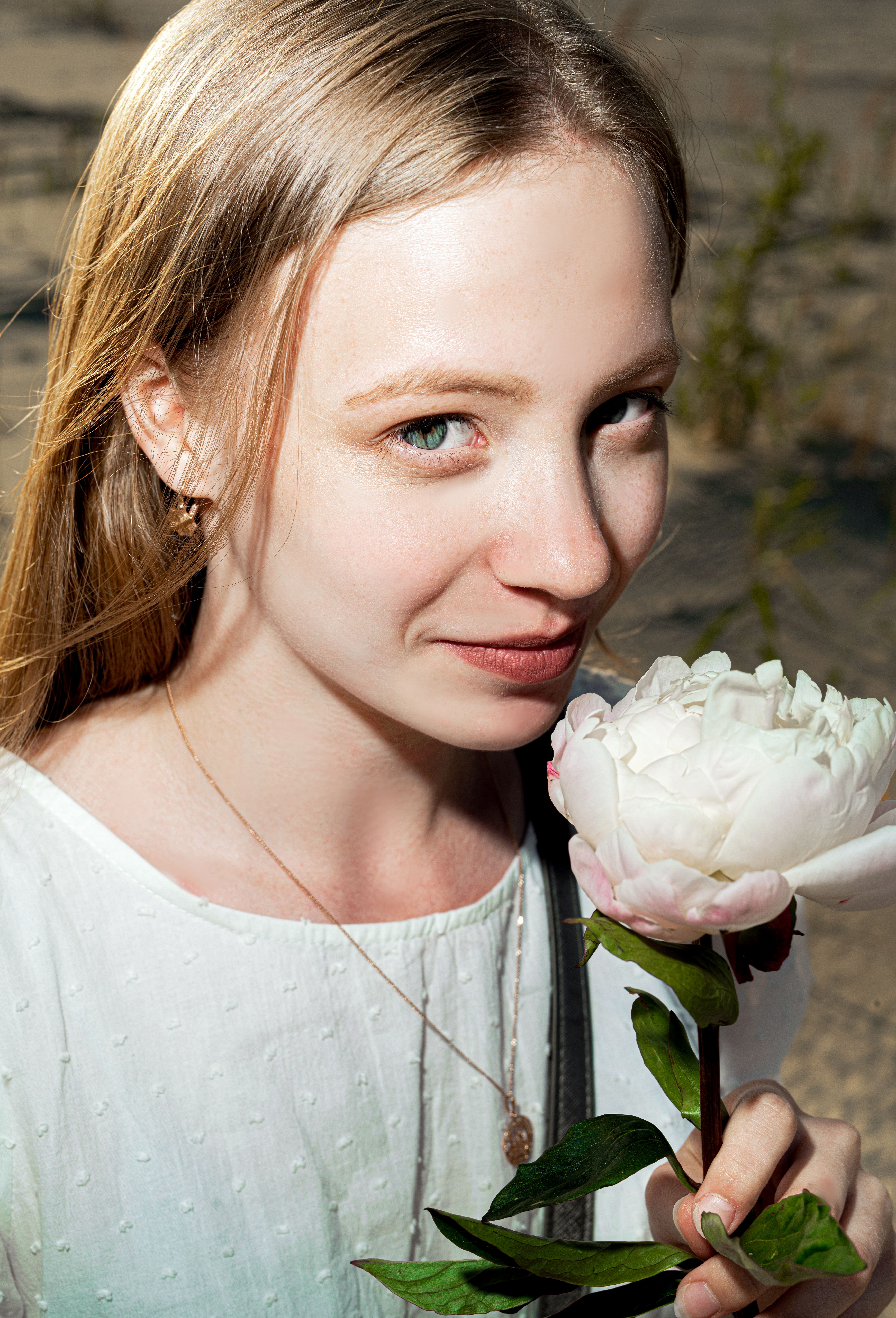 Blonde woman smiling while holding a white peony flower outdoors in natural light.
