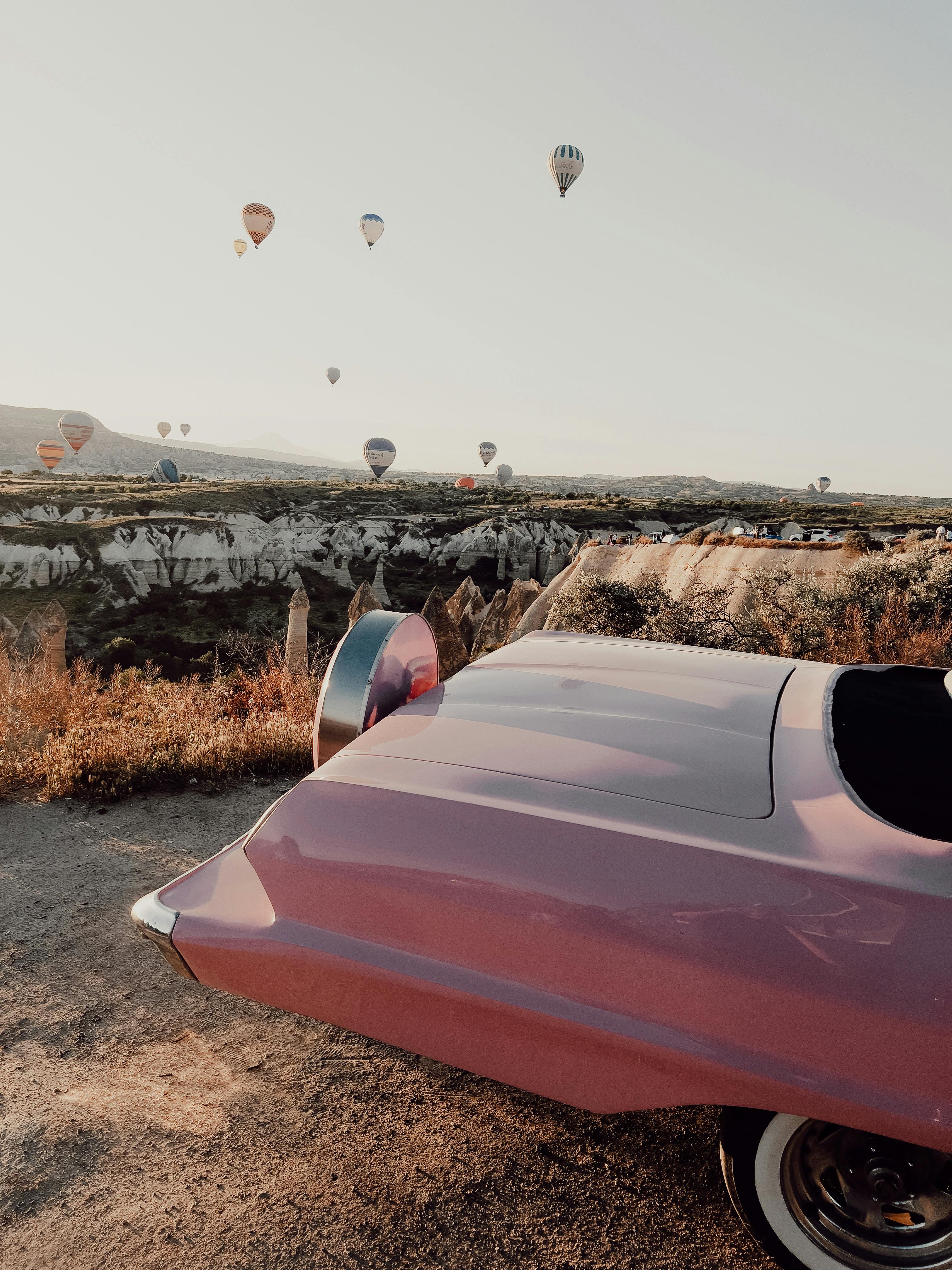 Scenic view of hot air balloons over Cappadocia landscape with pink convertible in the foreground.