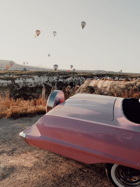 Scenic view of hot air balloons over Cappadocia landscape with pink convertible in the foreground.