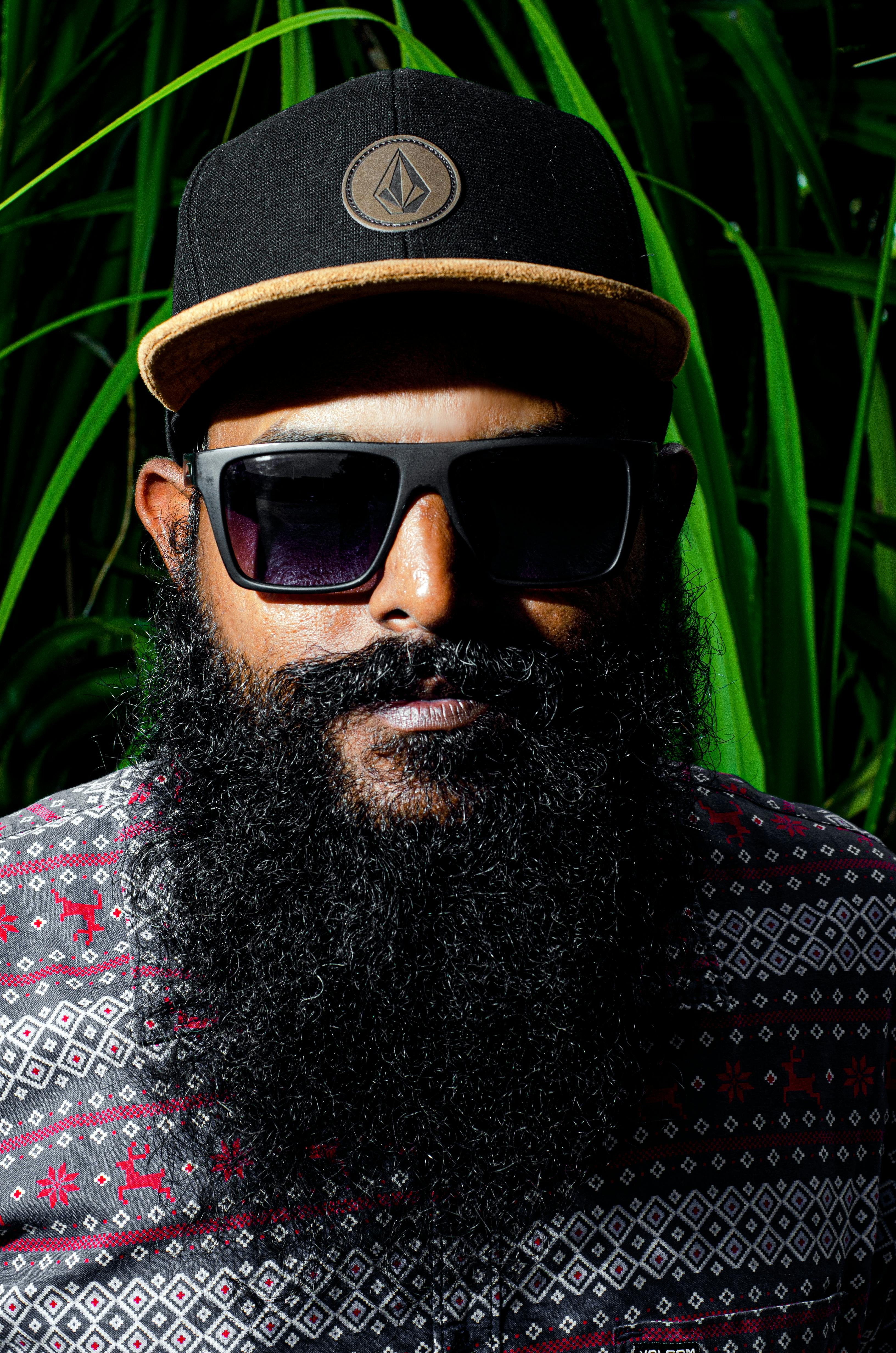 Portrait of a stylish man with a beard wearing sunglasses and cap against lush green background.