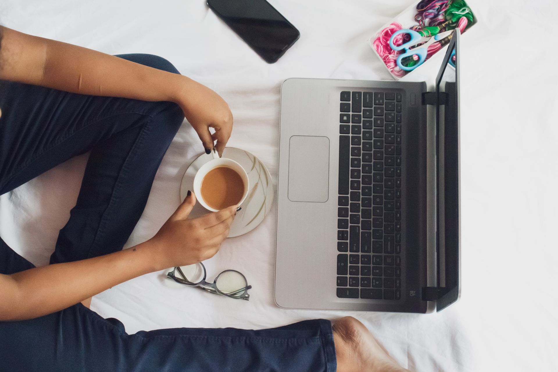 Comfortable remote work setup featuring a laptop, coffee, and essential tools on a white bed.