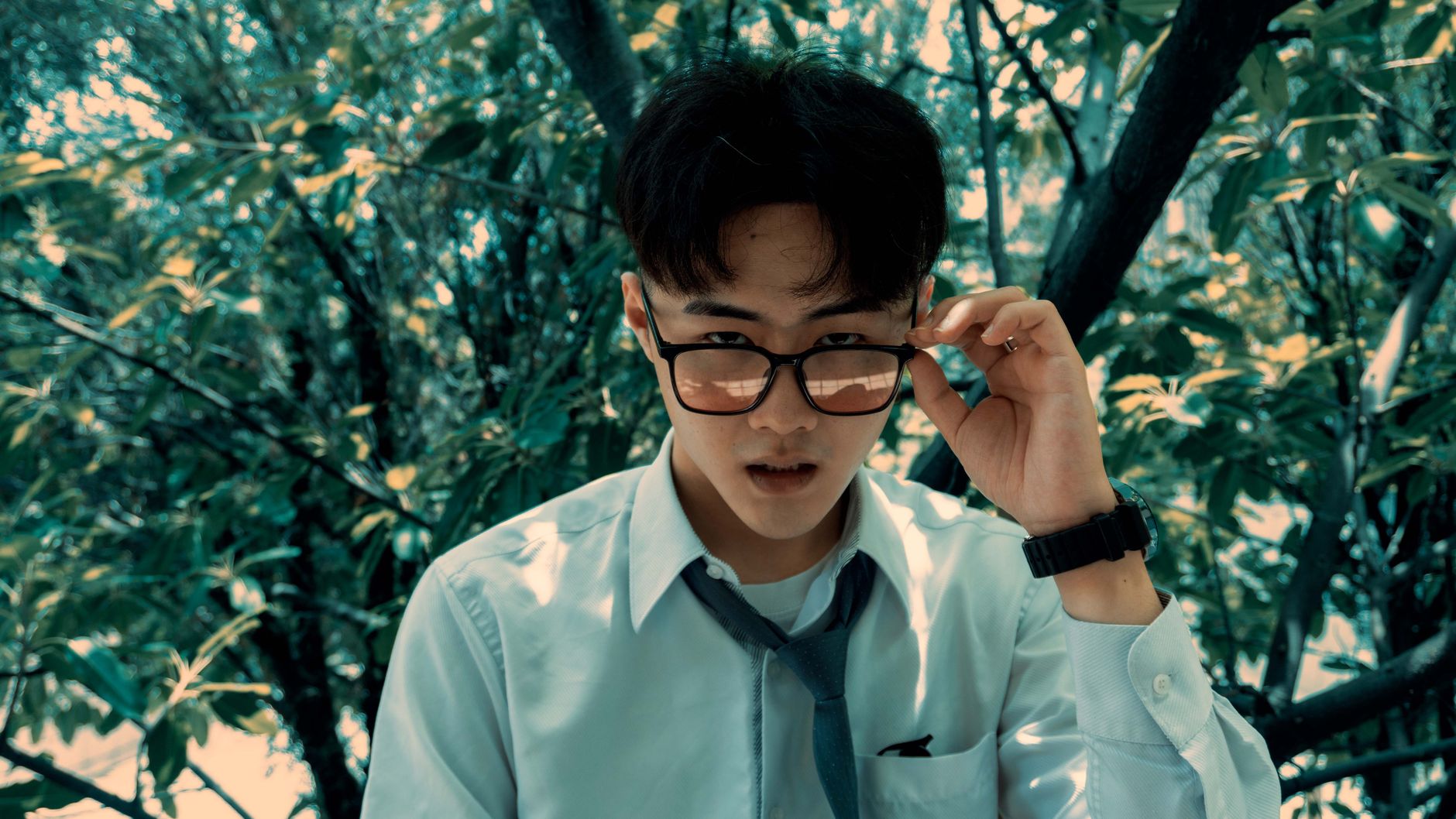 Portrait of a young man with glasses in a white shirt and tie outdoors among green trees.
