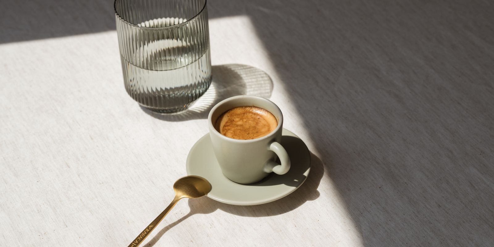 A minimalist setup with a coffee cup and glass of water in warm sunlight, featuring a golden teaspoon.