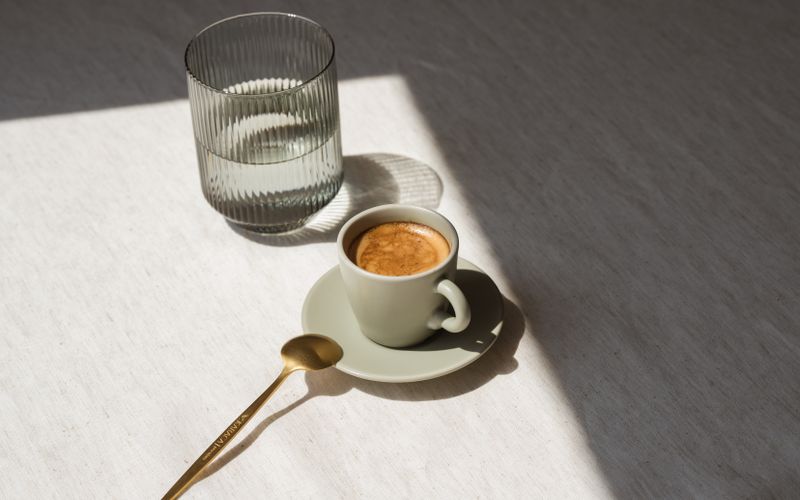 A minimalist setup with a coffee cup and glass of water in warm sunlight, featuring a golden teaspoon.