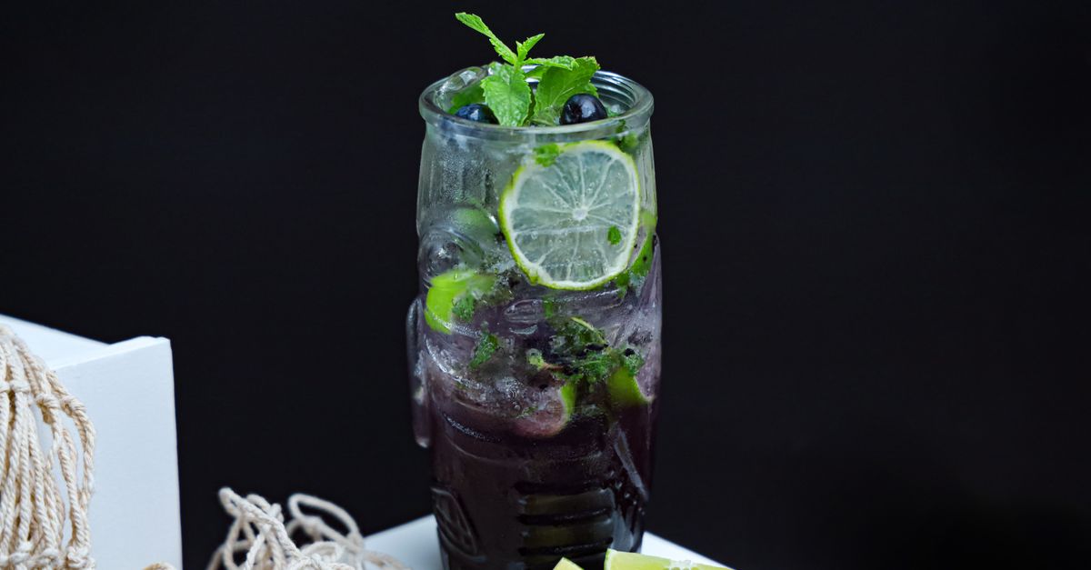A glass of dark purple mocktail with ice and a lemon wedge, sitting on a nightstand next to a book and a sleep mask