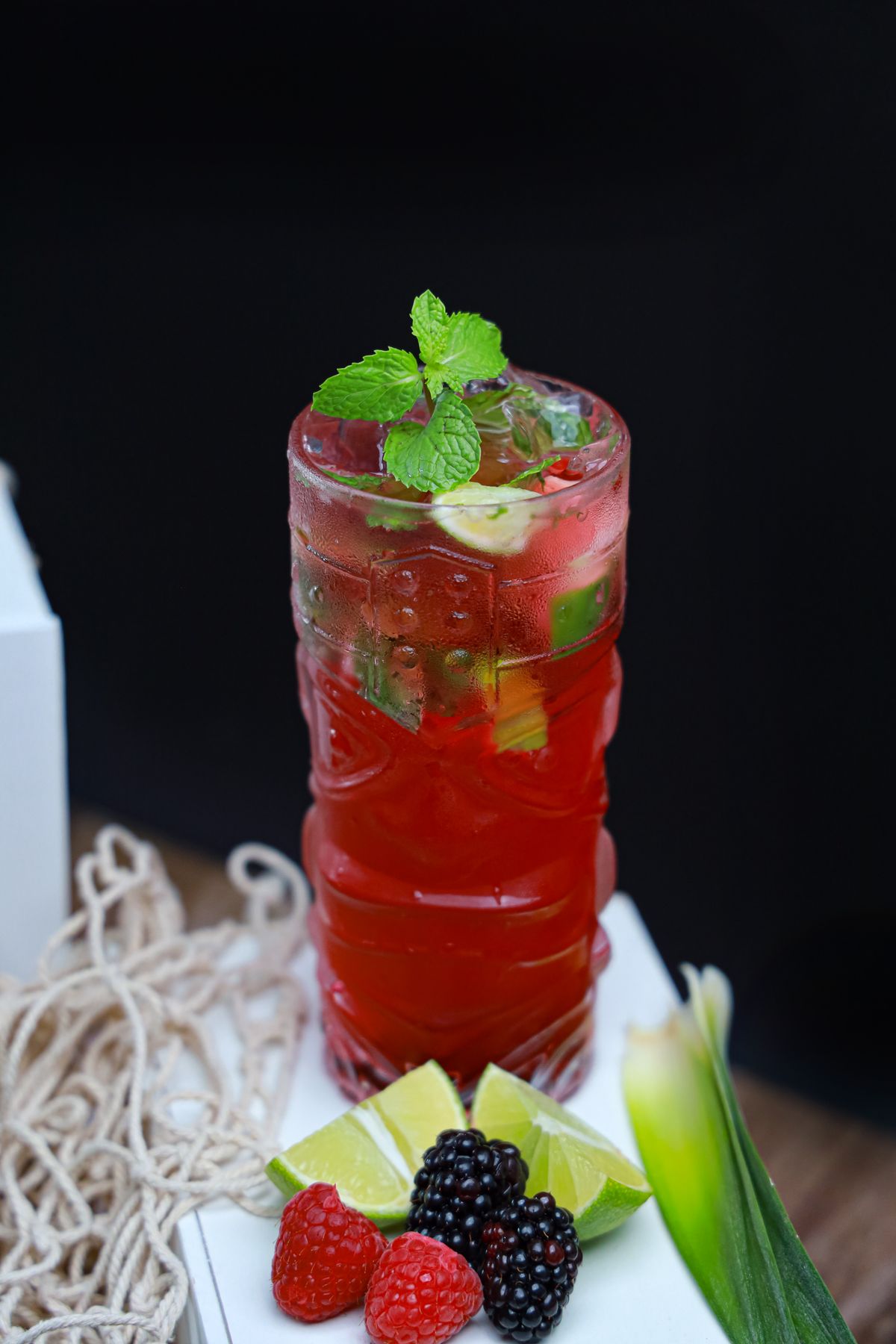 Tall red berry mojito with mint and lime in a textured glass beside raspberries and blackberries