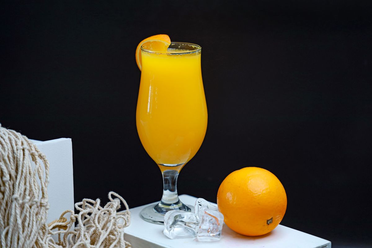Bright orange juice in a hurricane glass with an orange peel garnish beside a whole orange