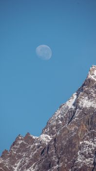 Serene view of the moon above a rugged, snow-covered mountain peak under a clear blue sky.