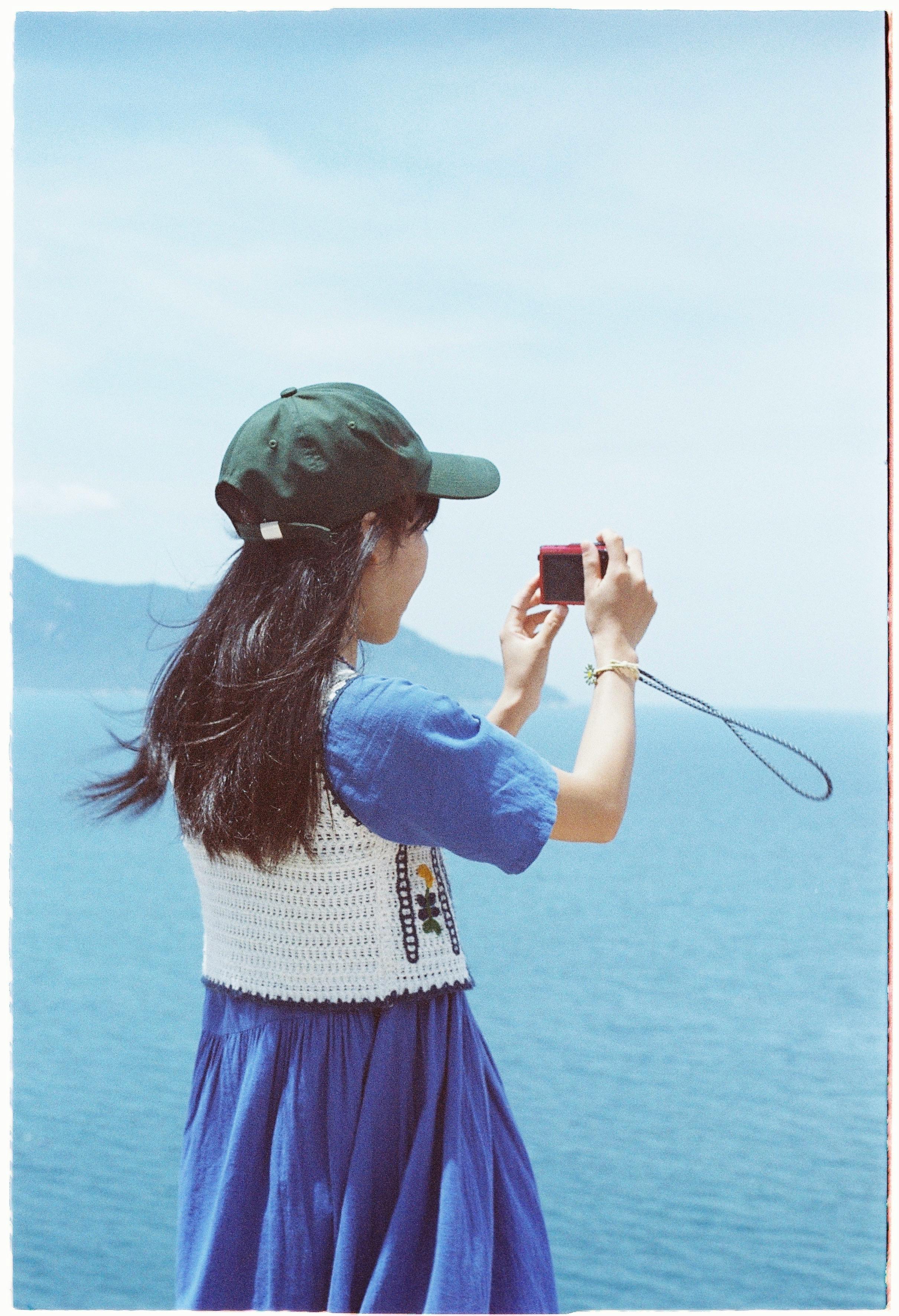 Woman in blue dress takes photo with vintage camera by the sea under a clear sky.