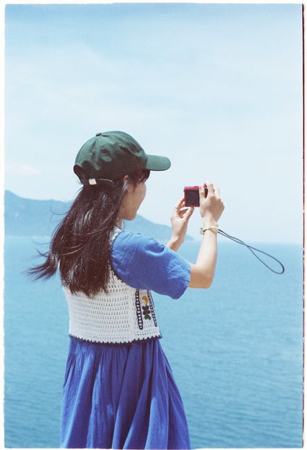 Woman in blue dress takes photo with vintage camera by the sea under a clear sky.