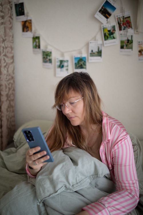 Free Woman in pink striped pajamas using smartphone in cozy bedroom setting featuring hanging photos. Stock Photo