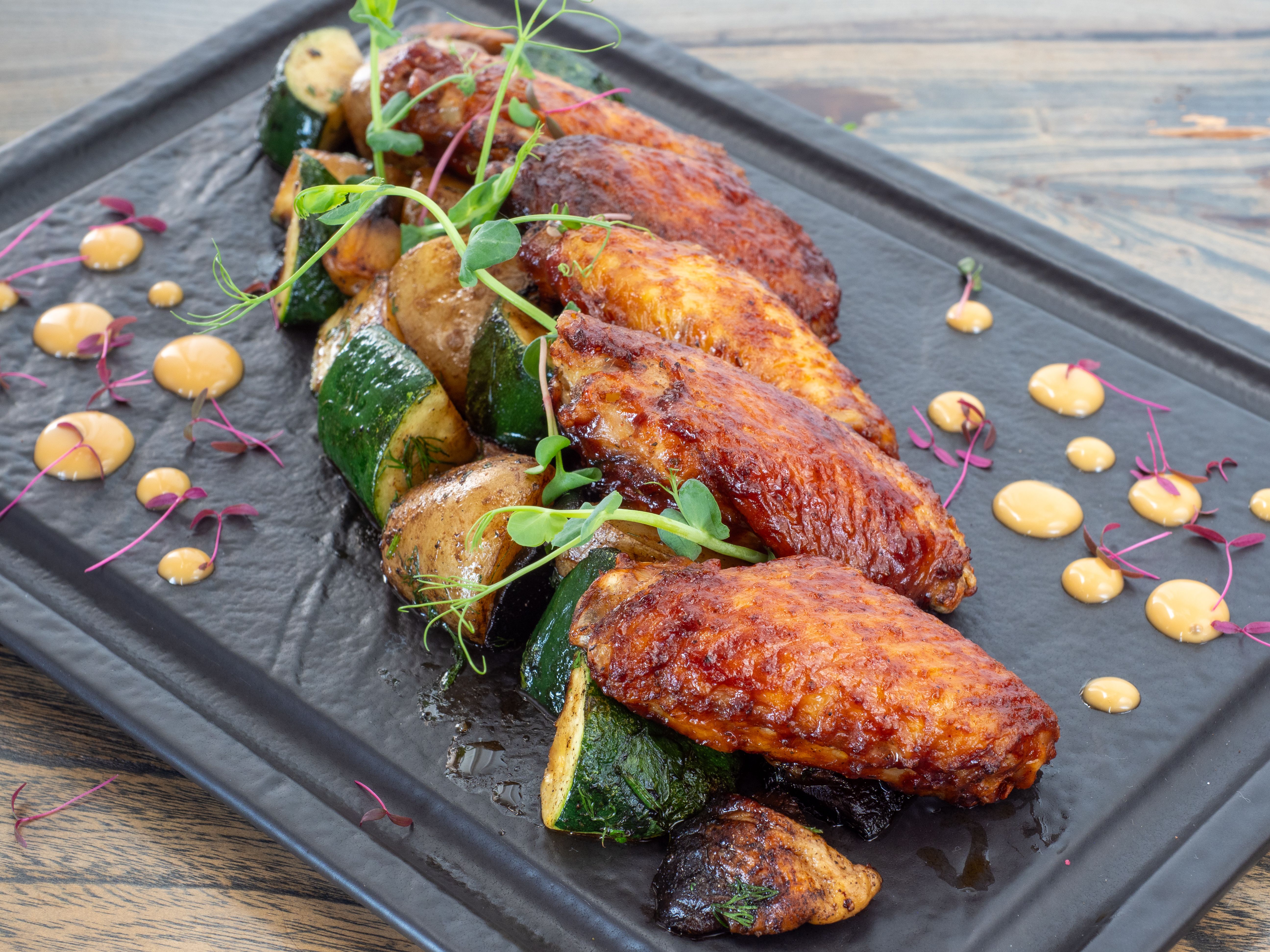 Close-up of grilled chicken wings with roasted vegetables on a black tray.