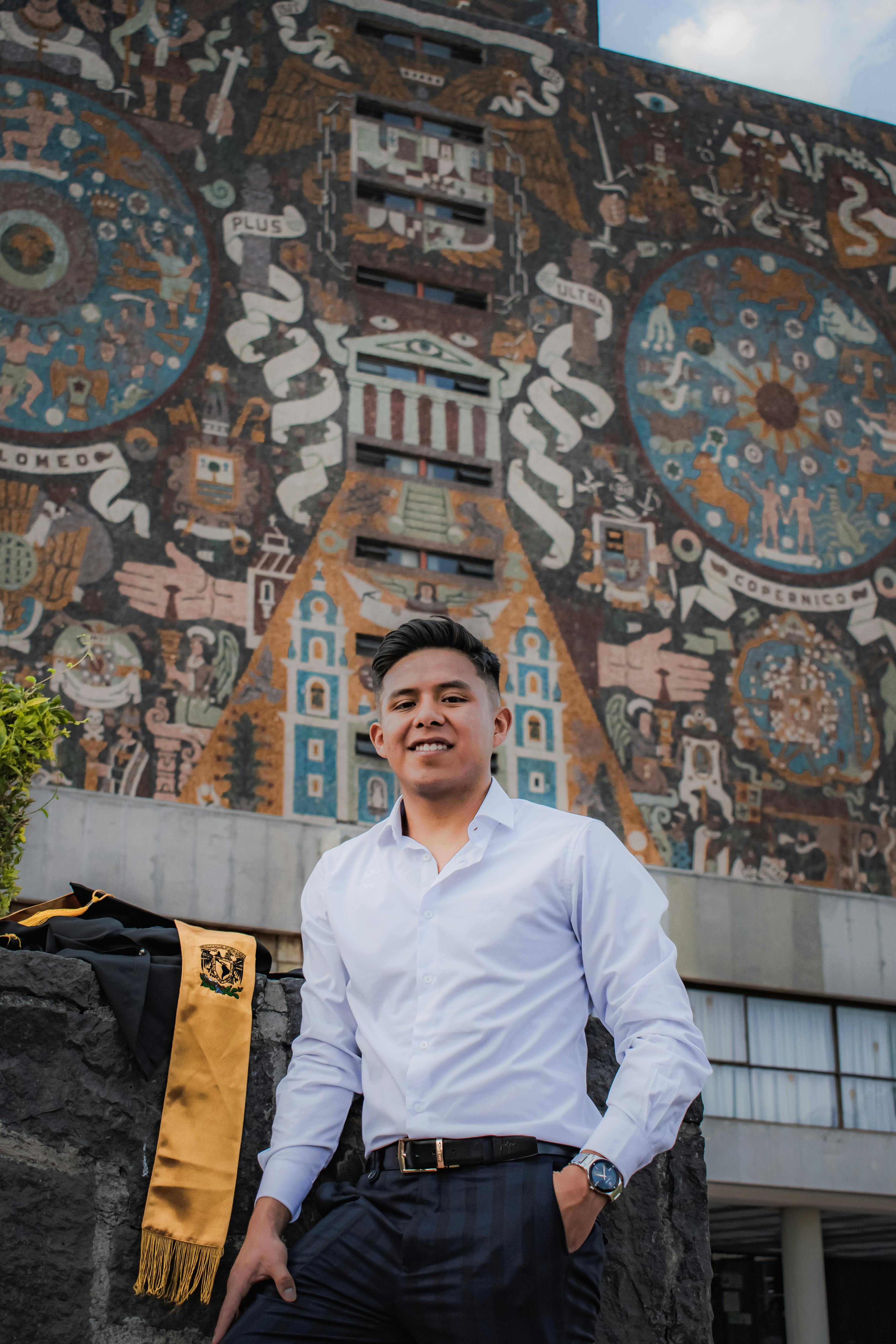 A young man in a white shirt stands proudly in front of the iconic UNAM library mural in Mexico City.